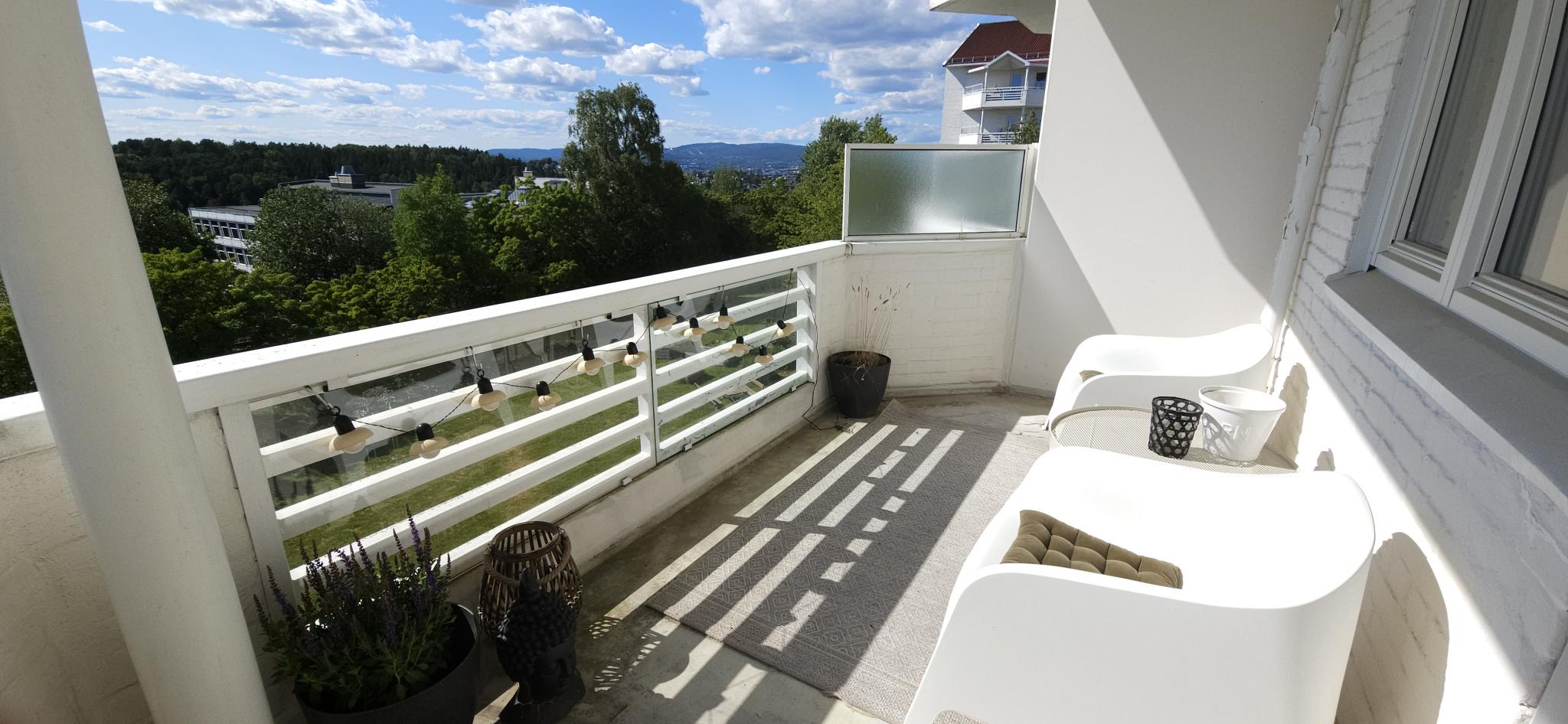Romslig solrik balkong med utsikt mot grøntområde, byen og Holmenkollen og Grefsenkollen. Solnedgang mellom Hk og Gk.
