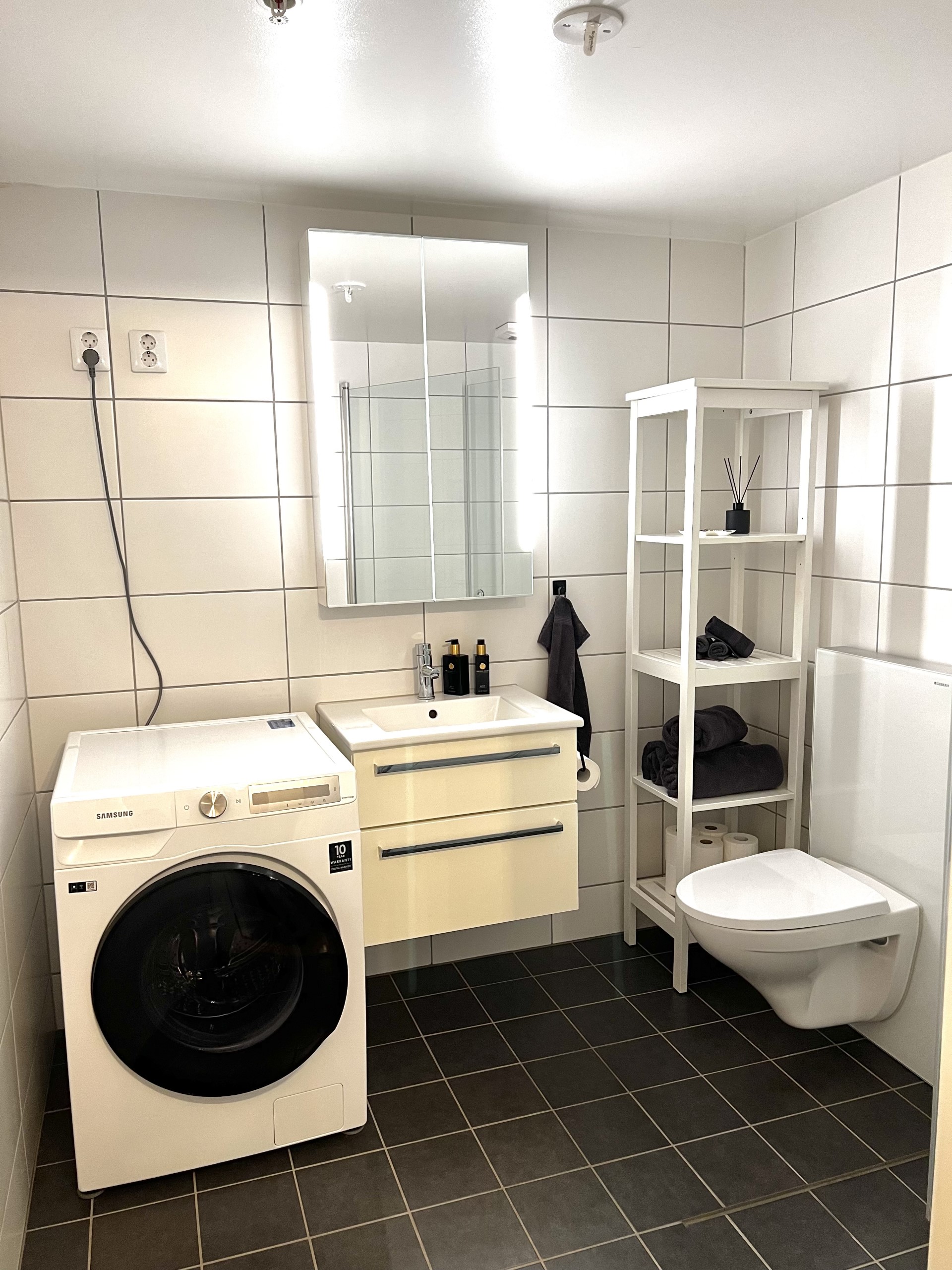 Bad med vegghengt toalett, servant med skuffer og hyller i speilskap. Kombinert vaskemaskin og tørketrommel som er inkludert i husleien. 