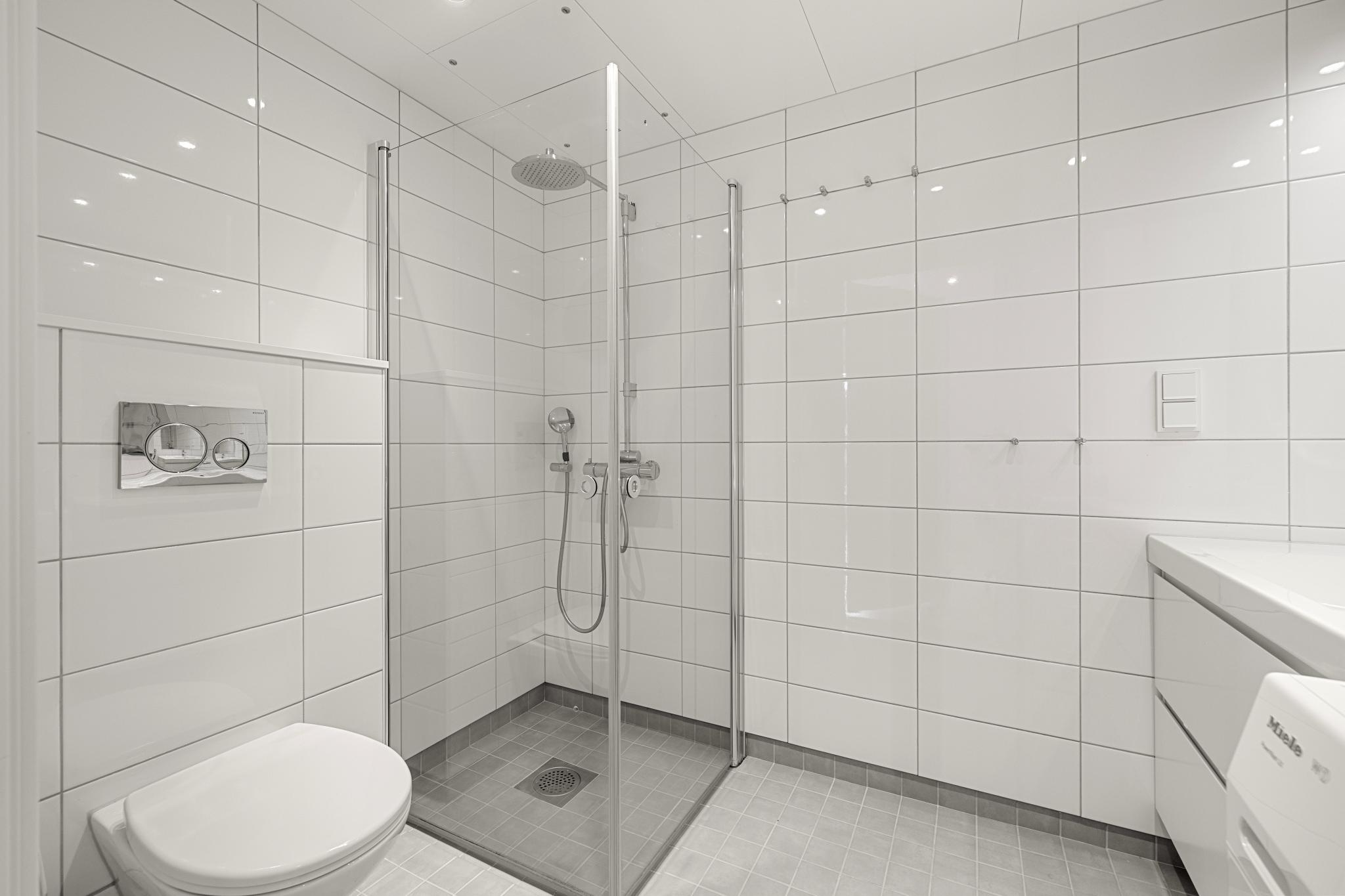 Badet er utstyrt med vegghengt toalett, servant med skuffer og dusjnisje med innfellbare glassdører. 