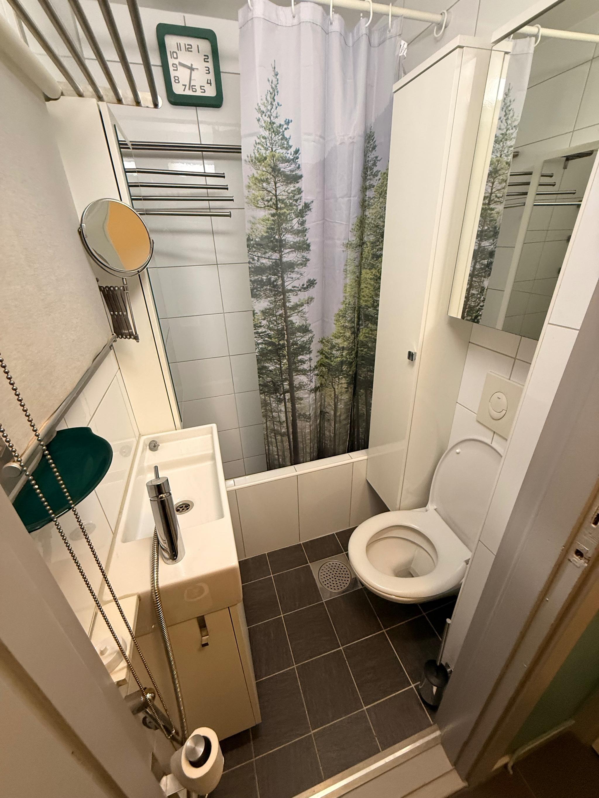 Lyst og kompakt bad med dusj, toalett og servantskap. Fliser på gulv med varme og vegger, samt håndkletørker og speilskap for oppbevaring.