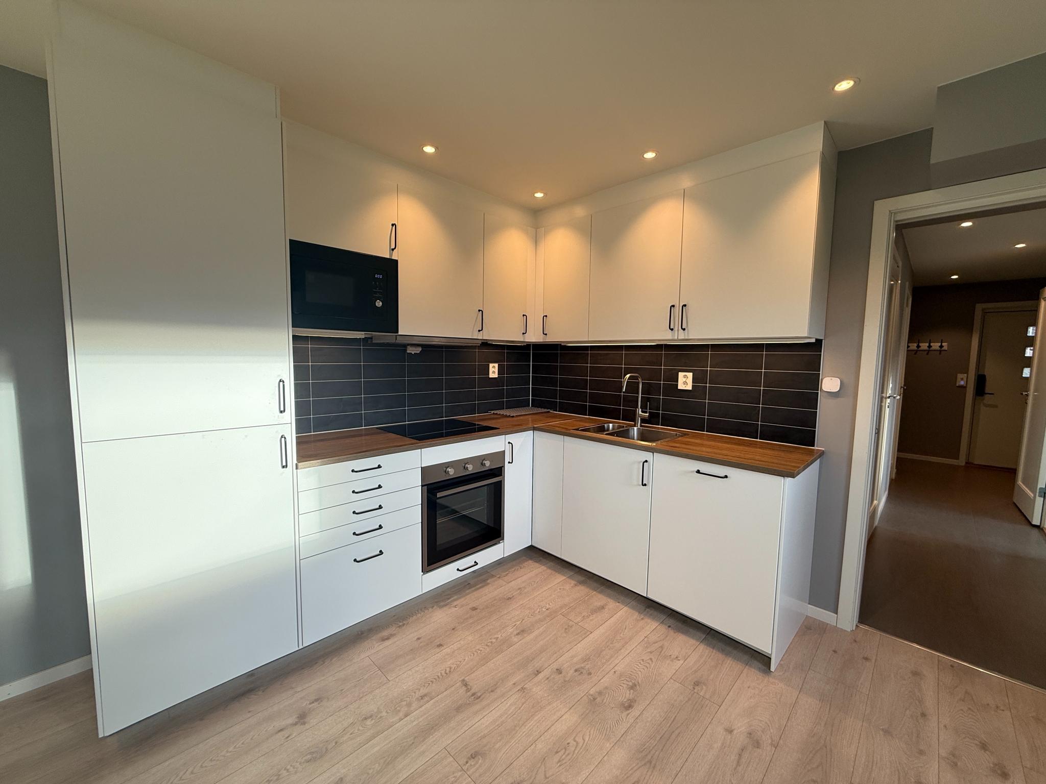 Helhetlig bilde av kjøkkenløsningen i L-form, med hvite fronter, mørk flis og benkeplate i trelook. God skapplass både over og under benk.