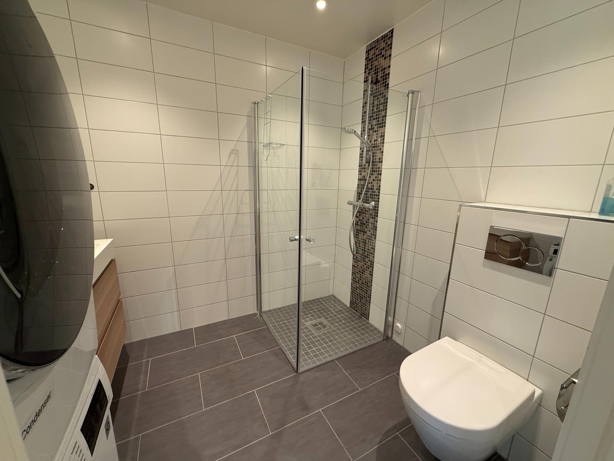 Oversiktsbilde av komplett bad med dusjhjørne i glass, vegghengt toalett og stilrene fliser på vegger og gulv.