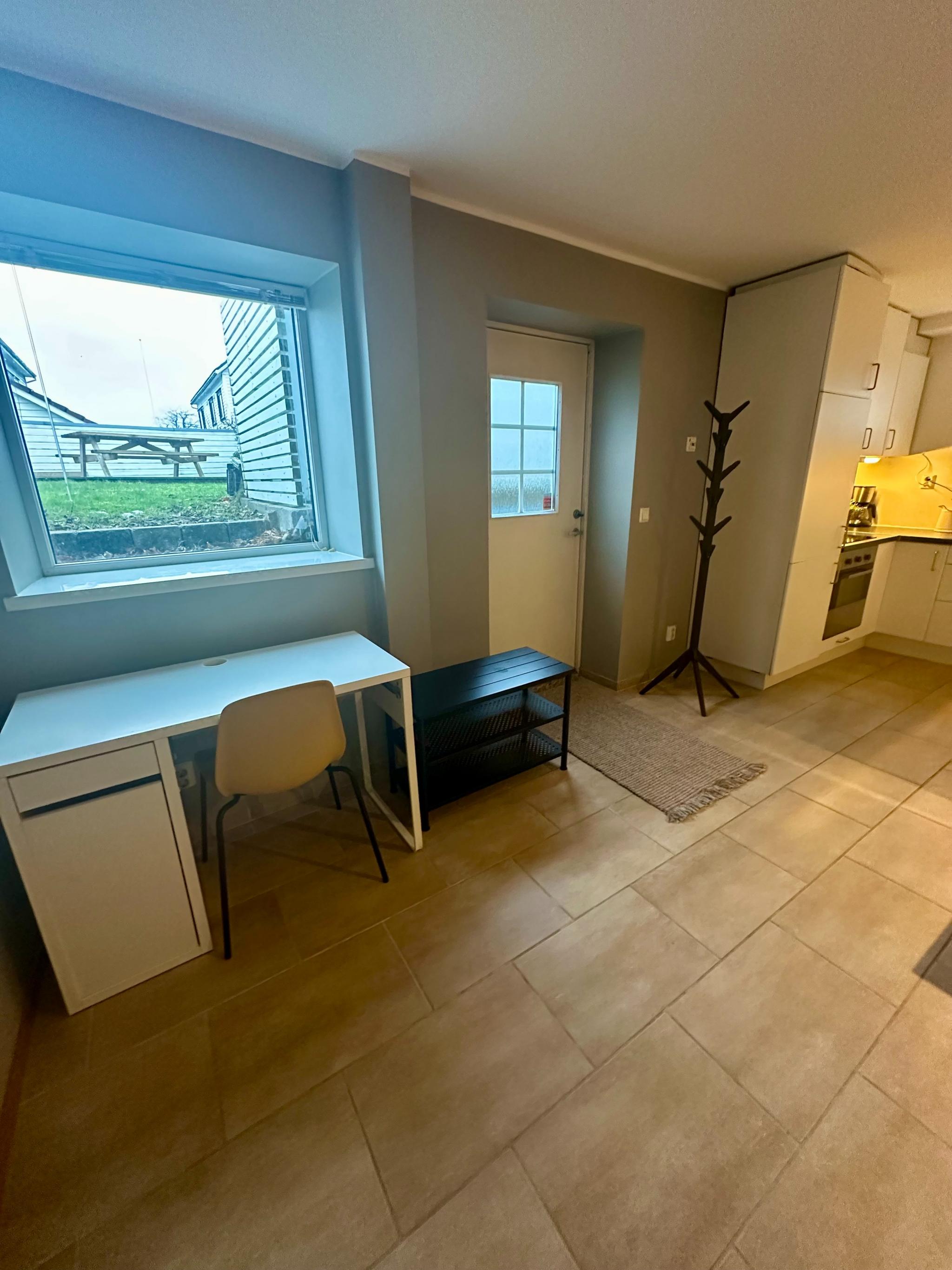 Inngangsdør, gang og et hjørne med skrivebord og stol. Vindu slipper lys inn og vender ut mot baksiden av huset/fellesarealer for boligen. 