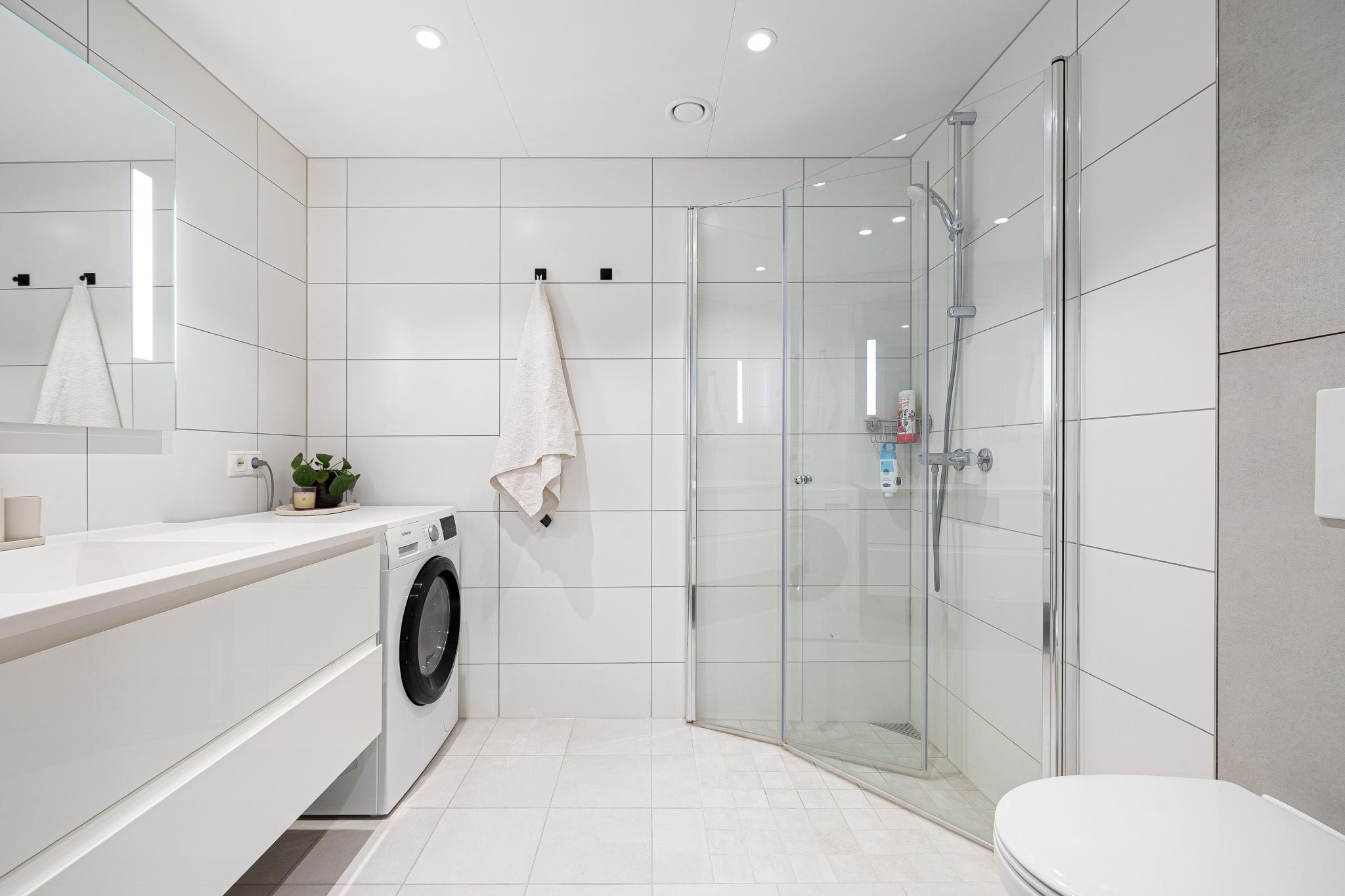 Flislagt baderom med moderne innredning, dusjhjørne og opplegg for vaskemaskin. Varmekabler i gulv. 